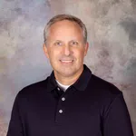 Smiling middle-aged man wearing a black polo shirt against a soft, mottled gray and beige background.