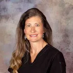 Woman with long wavy hair wearing black medical scrubs and silver earrings smiling in front of a neutral backdrop