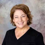 Smiling woman with short brown hair wearing black scrubs and gold jewelry against a neutral background
