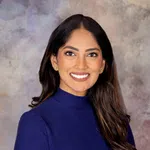 Smiling woman with long dark hair wearing a blue turtleneck against gray patterned background.