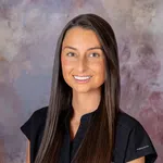 Young woman with long brown hair wearing a black shirt smiles against a soft purple and gray background.