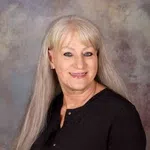 Smiling older woman with long blonde hair wearing a black blouse against a mottled gray background
