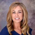 Smiling woman with long blonde hair wearing a blue top against a blurred gray and purple background
