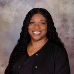 Smiling woman with long curly hair wearing a black top and jacket against a textured gray background
