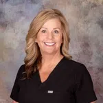 Smiling woman with long blonde hair wearing black medical scrubs against a textured background