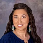 Smiling woman with long dark hair, wearing blue top and colorful dangling earrings against a blurred background.