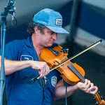 Musician wearing blue cap and shirt playing a violin on stage under a microphone.