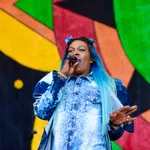 Female singer with blue ombre hair performing on stage with colorful abstract background art.