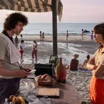 Young man and woman talking at a beachside stand with bread, drinks, and people walking along the shore at sunset.