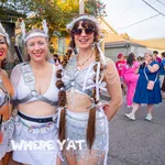 Three women in futuristic silver festival costumes posing outdoors at a sunny street event with a festive crowd.