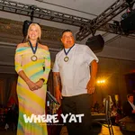 Two award recipients wearing medals stand on stage at a formal event with audience in background.