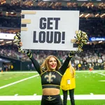 Cheerleader in New Orleans Saints uniform holding a GET LOUD! sign on football field with crowd behind