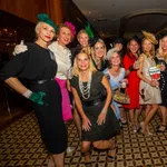 Group of women dressed in vintage-style dresses and hats posing indoors for a lively social event photo.