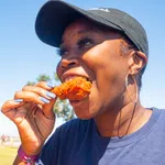 Smiling woman wearing a black cap and hoop earrings eating fried chicken outdoors on a sunny day
