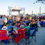 Crowd enjoying an outdoor music festival with people sitting on chairs and socializing near the stage at sunset.