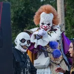 Three people in horror costumes including a clown with red hair, a skull-faced figure, and a woman in a red and gold jacket.