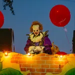 Costumed clown with red hair and white face paint holding red balloons at night during parade float event.