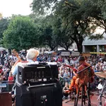 View from stage of a live band performing to a large outdoor crowd under trees at a music festival.