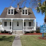 White historic house with a wraparound porch, manicured lawn, and a blue directional sign near a palm tree.