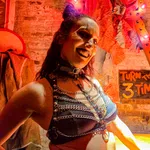 Woman in black leather harness and chains smiles against a brick wall with Halloween decor and overlaid text sign.