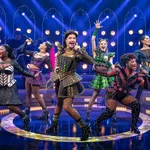 Six female performers sing and dance energetically on a brightly lit stage with colorful costumes and fishnet stockings.