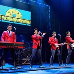 Four male performers in red jackets play instruments and sing on stage with musical notes backdrop in blue lighting.