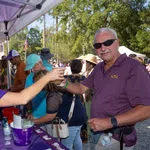 People enjoying a sunny outdoor festival, sharing drinks under a tent with vibrant purple banners.