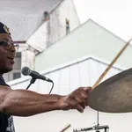 Man wearing a beret playing drums energetically at an outdoor performance with houses in the background