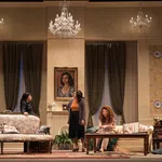 Theatrical stage scene with five women in vintage living room setting under chandeliers and art-adorned walls