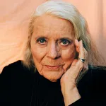 Elderly woman with silver hair, wearing black, resting her face on her hand against a soft peach background.