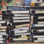 Stack of vintage VHS tapes with handwritten labels on a cluttered desk surrounded by posters and household items.