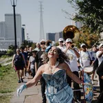 Woman in blue corset and gloves dances joyfully in front of a marching band and parade crowd on sunny day.