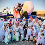 Group of people in white costumes and wings posing outdoors at a festival with a ferris wheel and colorful float.
