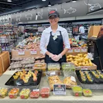 Smiling store employee presenting various homemade dips and salsas at a grocery store sampling table.