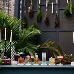 Outdoor bar setup with assorted cocktails, lit candles, greenery, and dried herbs hanging on a dark backdrop.