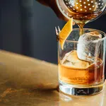 Pouring an amber cocktail over a large ice cube in a glass with a lemon peel garnish clipped to the rim