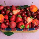 Basket filled with fresh strawberries, cherries, peaches, and apricots on a pink surface