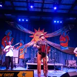 Live band performing on stage with accordion, guitar, and bass under bright lights and colorful sun backdrop