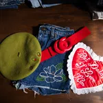 Green beret, denim shorts with floral embroidery, red belt, and heart-shaped pillow with love message on wooden surface.