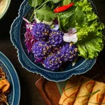 Colorful Thai dishes including purple flower-shaped dumplings, spring rolls, pad thai, and curry on ornate plates.