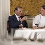 Two men smiling and toasting with red wine in an elegant dining room with gold wallpaper and exposed brick.