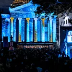 Crowd at night watching blue and gold light projections on a classical building during Luna Fête festival.