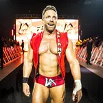 Muscular male wrestler in red trunks and vest walking confidently toward ring with audience and lights behind him