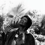 Black and white photo of a woman looking up surrounded by large palm leaves in soft light.