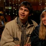 Teen boy and girl smiling and sitting together in a warmly lit indoor setting with people in the background