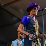 Musician in purple hat and vest singing into microphone while playing tambourine on stage at outdoor event.