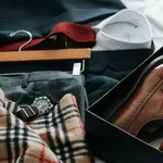Flat lay of men's brown dress shoes in a box, plaid scarf, gray trousers, watch, and a white shirt on a dark suit jacket.