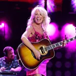 Female country singer in pink dress playing acoustic guitar on stage with vibrant purple lighting and smiling joyfully.