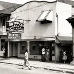 Black and white street scene with Dew Drop Cafe bar, cocktail lounge, hotel, and people walking and standing outside.