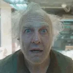 Older man with wild white hair and surprised expression looking directly at the camera indoors.